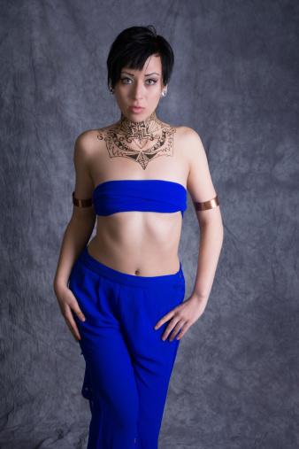 Vertical studio shot on gray of young woman in blue bandeau and pants with henna neck/chest piece.