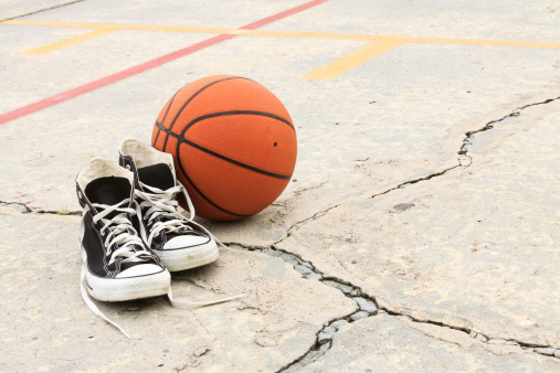 A pair of old fashioned athletic shoes and a basketball on the ground of a ghetto playground.
