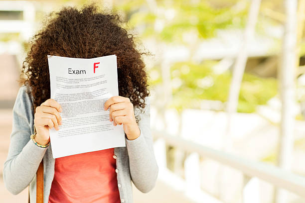 Disappointed Girl Holding Result In Front Of Face On Campus Disappointed teenage girl holding failed exam result with F grade in front of face on university campus. Horizontal shot. letter f photos stock pictures, royalty-free photos & images