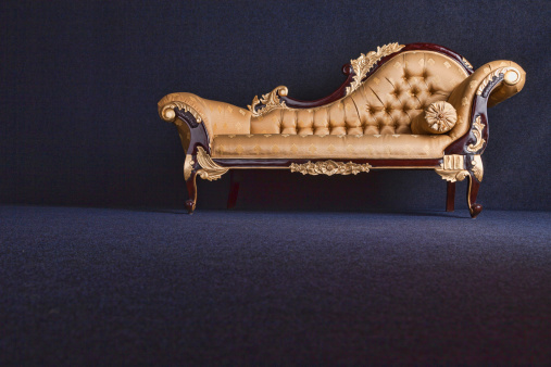 Low level view of a gold brocaded chaise longue in an empty, dark carpeted room.