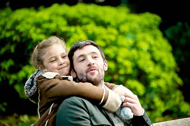 Young girl aged 6 to 7 years with mid adult man aged 30 t0 35 years, girl throwing her arms around her dad from behind, outdoors, autumn/ winter time