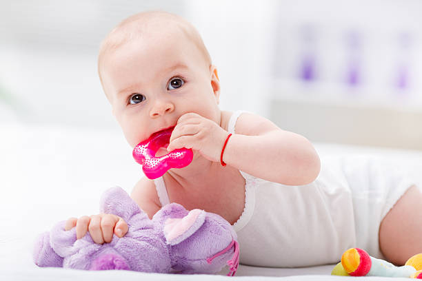 Baby chewing a teething toy on bed. Leaning on a teddy bear.