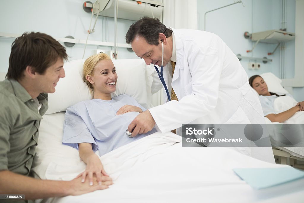 doctor hombre usando un estetoscopio a su sonriente mujer embarazada - Foto de stock de 20 a 29 años libre de derechos