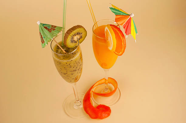 Kiwi smoothie and citrus juice in glasses decorated with citrus and kiwi fruit slices, paper umbrella, plastic straw and orange peel isolated on beige paper background. Selective focus.