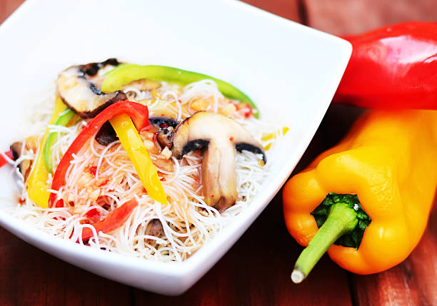 A delicious white bowl of vegetable stir fry noodles with yellow and red peppers besides the dish. Popular Chinese dish, healthy vegetarian option.