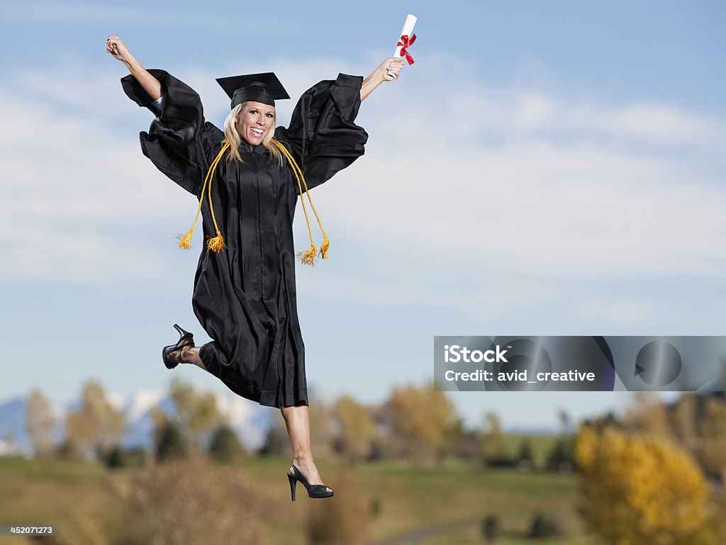 Jumping: Beautiful Female School Graduate A beautiful young caucasian woman wearing graduation gown and holding diploma certificate jumps with excitement. Mid air with sky and clouds in the background. Arms Raised Stock Photo