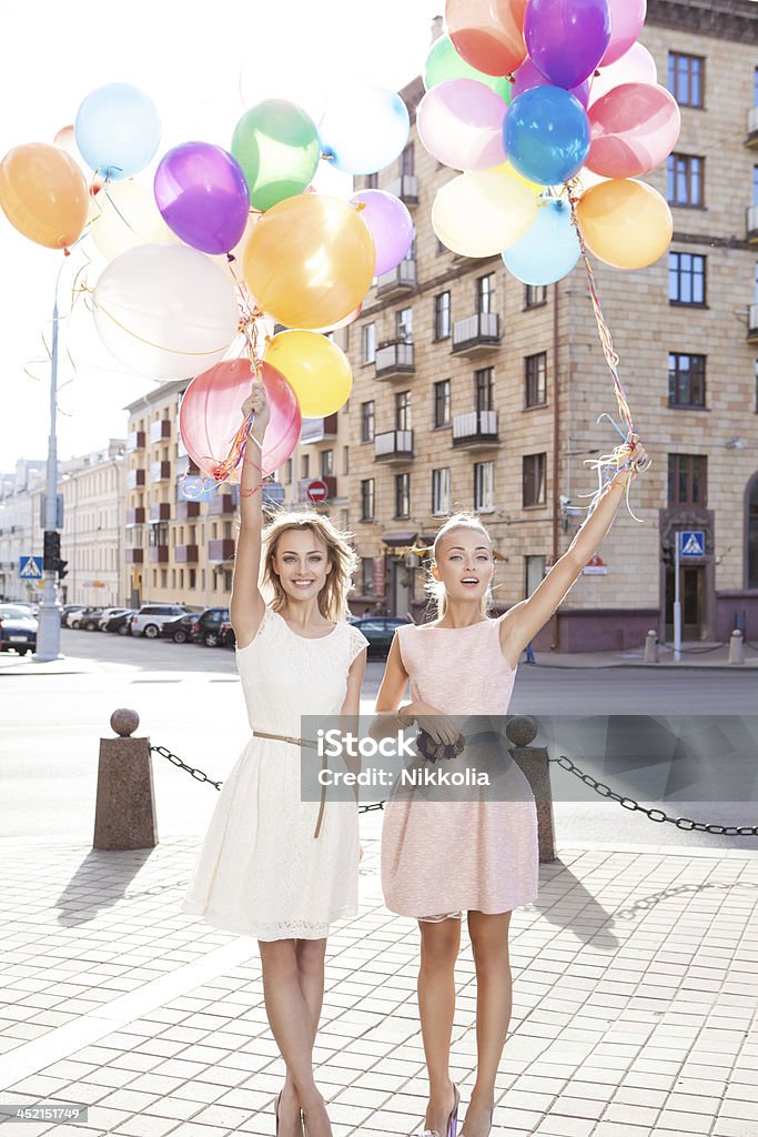 two beautiful ladys in retro outfit holding balloons two smiling girls in white and pink dresses holding bunches of multicoloured balloons in sun in the city street Adult Stock Photo