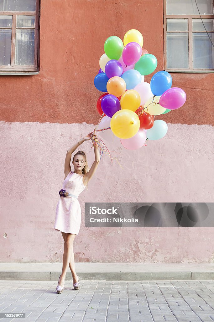 Young woman holding bunch of colorful balloons on street beautiful girl with closed eyes in pink dress  holds a bunch of balloons in front of old wall Adult Stock Photo