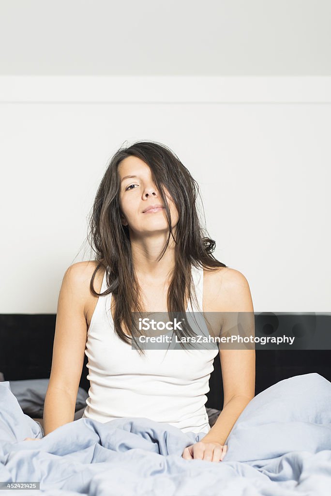 Tired young woman waking up after sleep Tired young woman waking up after a nights sleep giving the camera a lazy smile as she sits in her bed in her sleepwear trying to motivate herself, with copyspace Adult Stock Photo