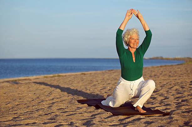 зрелая женщина, упражнения йоги на пляже — - senior adult outdoors wellbeing sky стоковые фото и изображения