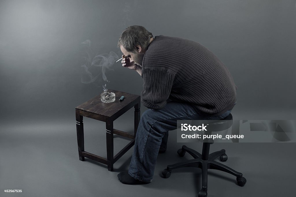 Depressed young man sitting on a chair, smoking cigarette A young caucasian man is alone, stressed, got fired from work is unemployed and sad. Addiction Stock Photo