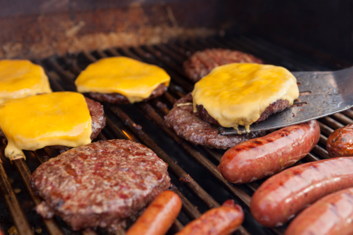Outdoor photo of a grill with hotdogs, sausages, hamburgers and cheeseburgers.  Shallow depth of field.  Focus is on the cheeseburger and spatula.