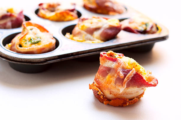 Mini Bacon Wrapped Omelet Bites A "Mini Bacon Wrapped Omelet Bite", with several more in the background.Made by wrapping a slice of bacon around inside of a mini-muffin tin and filing it with a mixture of scrambled eggs, chopped spinach, chopped onion, chopped tomato, sliced mushrooms, sausage crumbles and shredded cheese, then baking. muffin tin eggs stock pictures, royalty-free photos & images