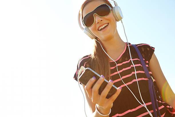 Side view of smiling modern middle age woman in white t-shirt and pink shorts listening to the music with headphones on the ocean shore.