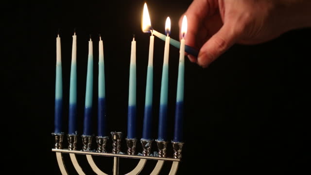 A menorah sits against a black background. A male hand enters from right and lights the candles.