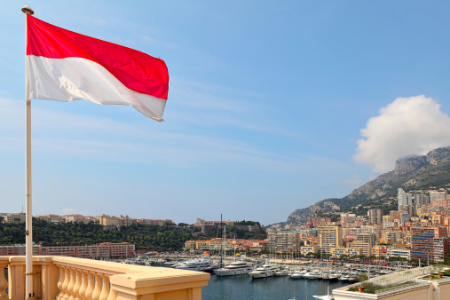 National flag, modern residential buildings and luxury yachts in Port Hercule under blue sky in Monte Carlo, Monaco.