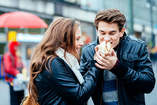 Eating a hot dog together Young couple eating a hot dog together in autumn - winter, small DOF, man at hot dog stand unsharp in the background. hot dog stand stock pictures, royalty-free photos & images