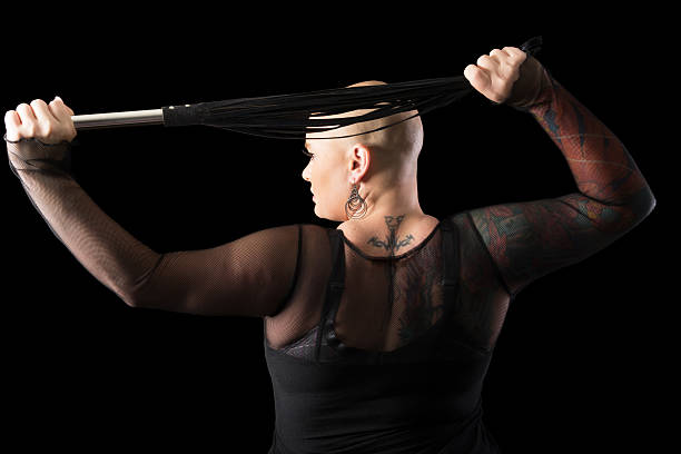 Shaved headed dominatrix holding flogger over head. Horizontal studio shot on black of young woman with shaved head holding black flogger behind head. dominatrix stock pictures, royalty-free photos & images