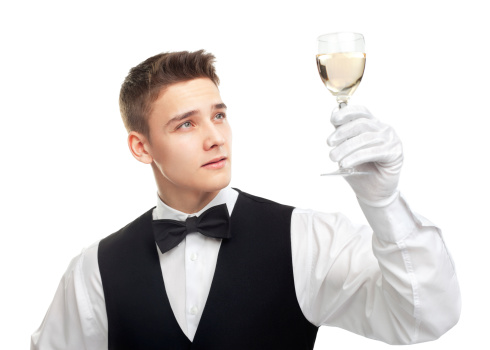 Portrait of young waiter looking at the glass filled with white wine isolated on white background