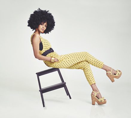 A young woman wearing a 70s retro jumpsuit while balancing on a stool in the studio