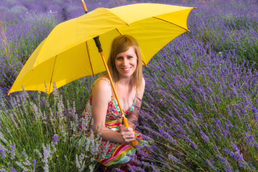 Young, attractive woman sitting in the lavender field holding open yellow umbrella,she has long blond hair, colorful summer dress and is smilling and looking to the camera.