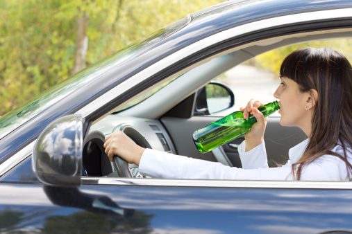 Inebriated female driver drinking alcohol directly from the bottle as she steers her car along the road posing a danger to others