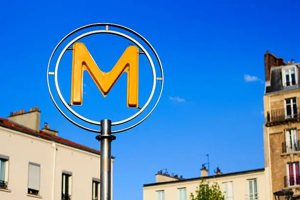 Parisian metro sign against blue sky with blurred building on the background, Paris, France