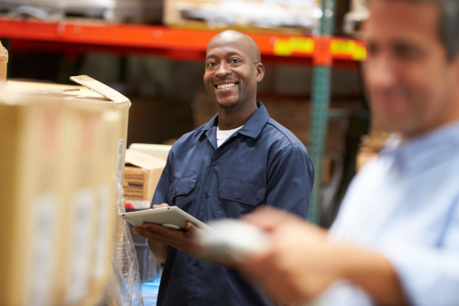 Manager In Warehouse With Worker Scanning Box In Foreground Smiling To Camera