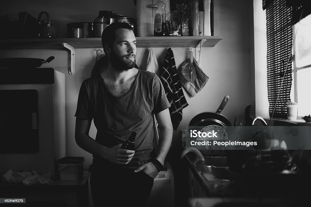 Giving thought to the day ahead A young man looking out of his kitchen window thoughtfully while enjoying a beer Home Interior Stock Photo