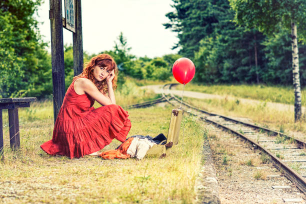 Teen girl in stylish beige dress sitting on vintage suitcase on abandoned railway in spring forest, lonely, looking away. Fashionable lady in retro image. Travel vacation concept. Copy text space