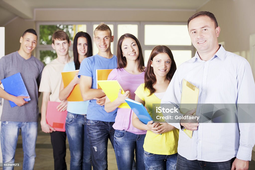 Teacher is standing in school hall with students. Teacher is standing in school hall and looking at the camera, his students is behind him. They are holding their notebooks. School Principal Stock Photo