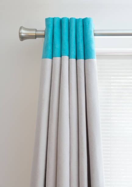 turquoise curtain tops a set of folded curtains on a stainless steel curtain rail. The tops are a in a vivid turquoise blue and light grey below. In the background is a grey wall and a closed roller blind in front of a bright window. curtain rail stock pictures, royalty-free photos & images