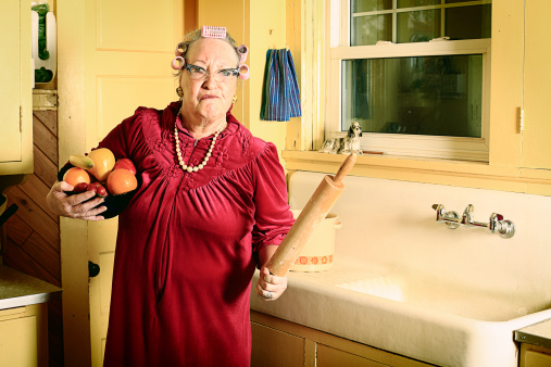 A grumpy granny in curlers and cat glasses stands with a bowl of plastic fruit and rolling pin in her kitchen near the sink. More granny images.
