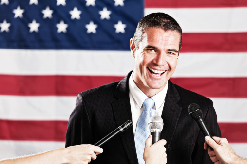 This handsome, confident-looking candidate, probably for political office, stands in front of the US flag gesturing and laughing in delight as he faces many microphones being held up eagerly by the Press.