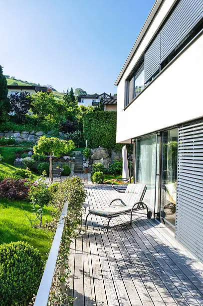 Outdoor view of a luxurious contemporary rectangular style house. Under blue sky. A large patio deck and deck chair invite to relaxation and sun bathing.  The terrace is right next to a beautiful garden. Vertical orientation. The size of the image amounts to 2275 px × 3425 px