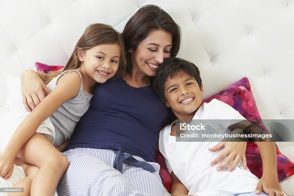 Mother And Children Relaxing In Bed Wearing Pajamas Mother And Children Relaxing In Bed Wearing Pajamas With Arms Around Children Smiling Family with Two Children Stock Photo