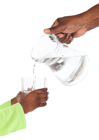 Hand of an african adult man is holding a jug of water and pouring it into a clear glass in the hands of a small african child with green sleeves.