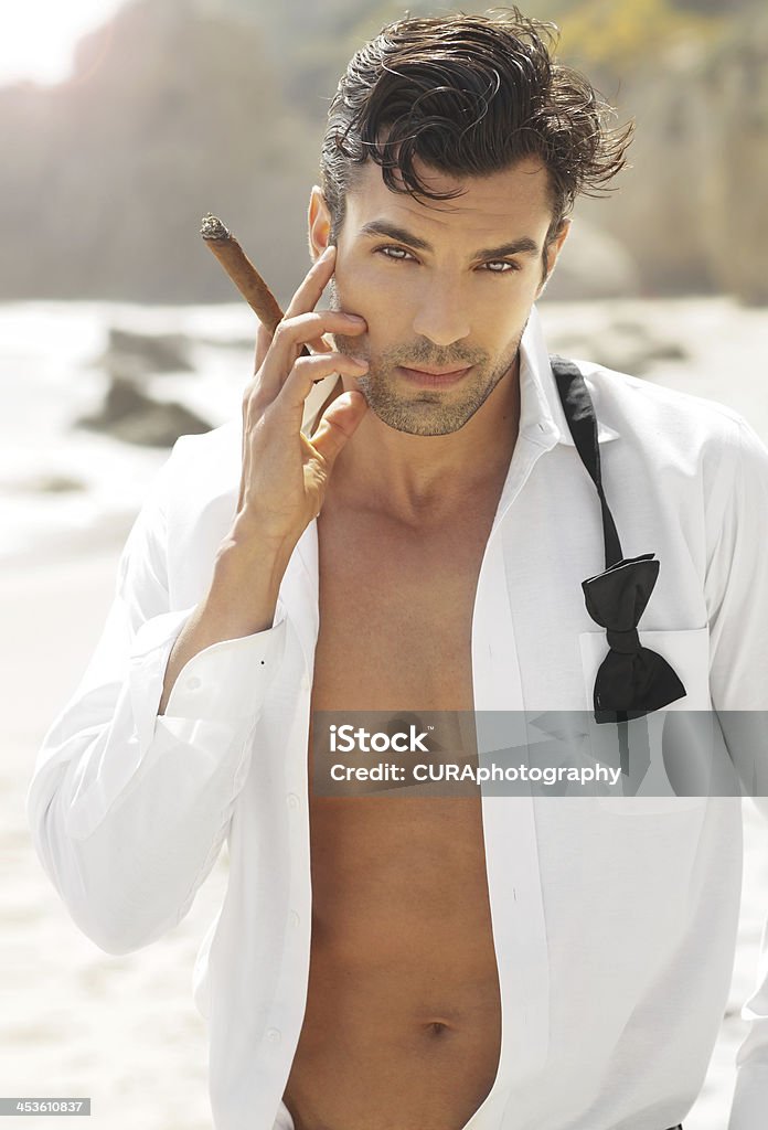 Sexy cool guy Great looking sexy male model with open white shirt and loose bow tie smoking cigar on beach Cigar Stock Photo