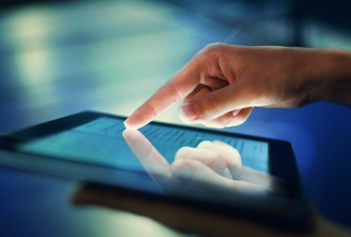 A finger touching the screen of a digital tablet.  In the foreground, a softly focused hand supports the device from below.  The hand that is touching the screen is sharply reflected in its glass.  The background is blurred and blue.