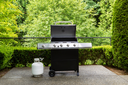 Horizontal photo of large barbeque cooker, with lid up, on concrete outdoor patio with woods background