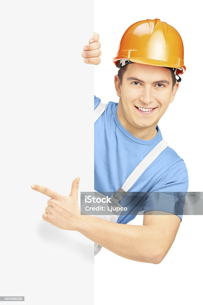 Young smiling construction worker with helmet on a panel Young smiling construction worker with helmet pointing on a blank panel isolated on white background Adult Stock Photo