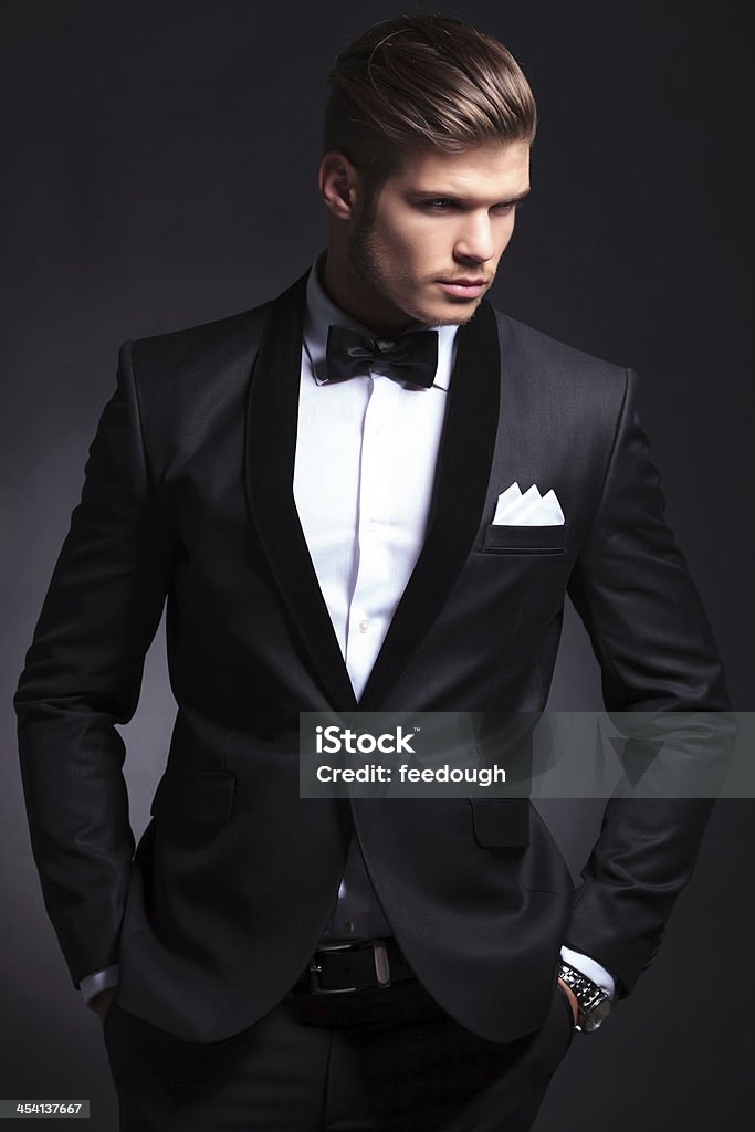 business man looks away waist-up picture of an elegant young fashion man in tuxedo looking away from the camera while holding hands in pockets.on black background Adult Stock Photo