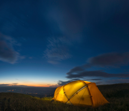 Warmly illuminated mountaineering dome tent pitched high on a grassy mountain below chrome blue dusk skies and overlooking the lights in the valley far below. ProPhoto RGB profile for maximum color fidelity and gamut.