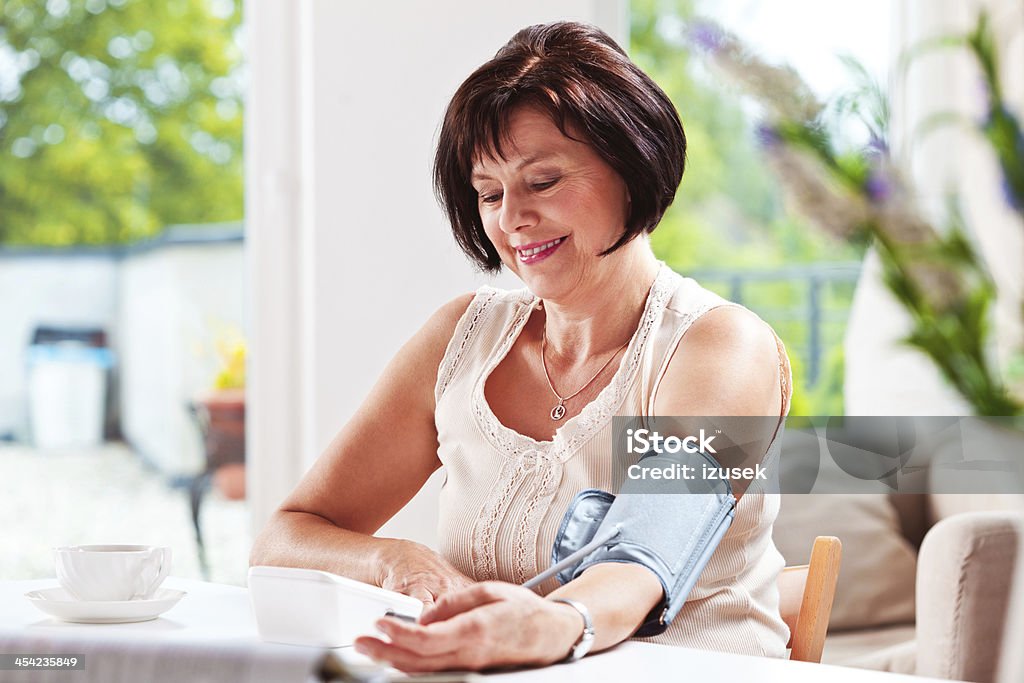 Woman checking blood presure Cheerful mature woman sitting at the table at home and checking her blood presure. Blood Pressure Gauge Stock Photo