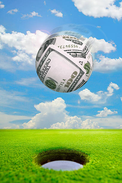 Golf ball made of money about to drop in the hole on greens stock photo