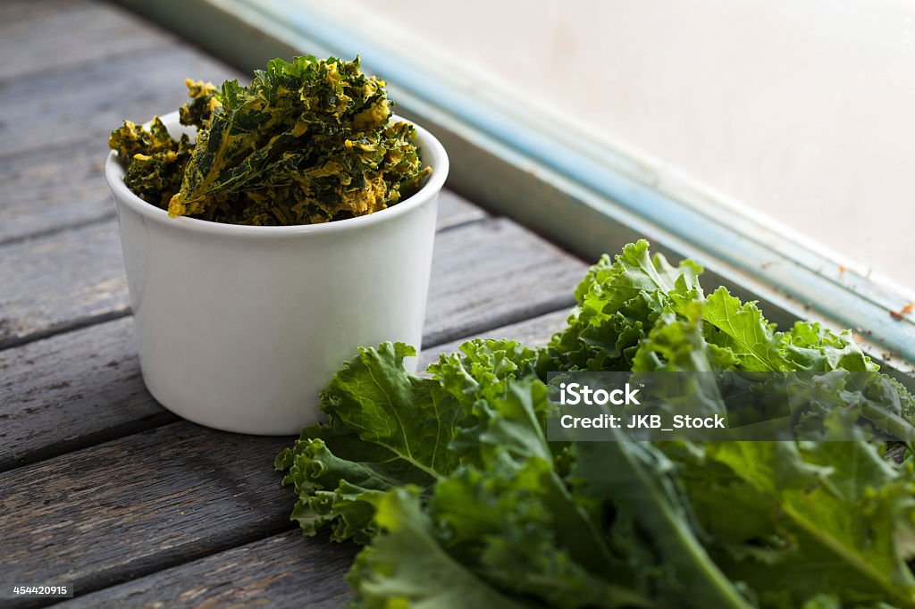 White bowl of kale chips next to fresh kale by the window Organic dehydrated kale chips in white bowl next to a bunch of fresh kale Agriculture Stock Photo