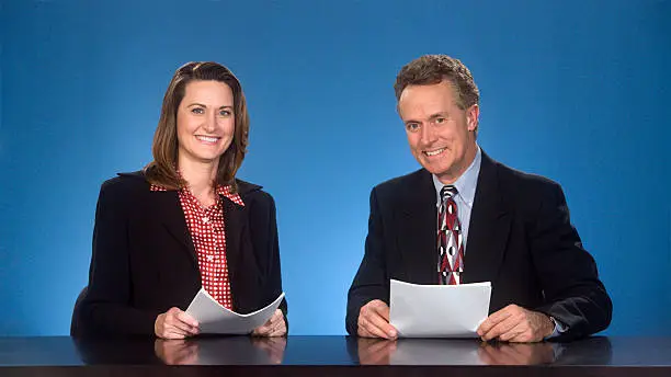 Male and female newcasters sitting at desk smiling at viewer.