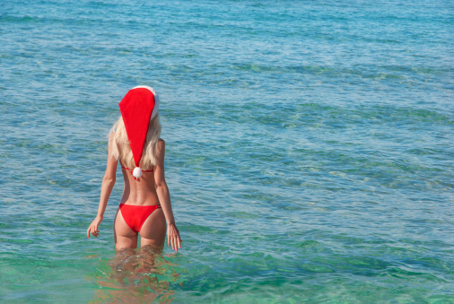 Beautiful young blonde woman in red christmas hat on sea beach. New year holidays in hot countries concept.