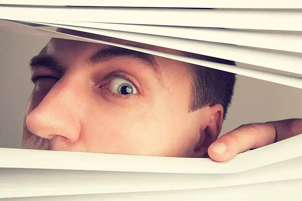 A young man looking through window blinds