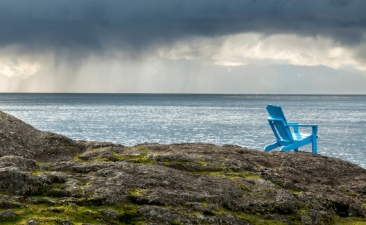 A blue Adirondack chair sits on the rocks overlooking the ocean as a rain cloud approaches.